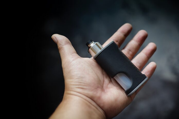 high end unregulated bottom feed squonk box mods with rebuildable dripping atomizer in hand on dark grey texture background, vaping device, vape gear, vaporizer equipment, selective focus